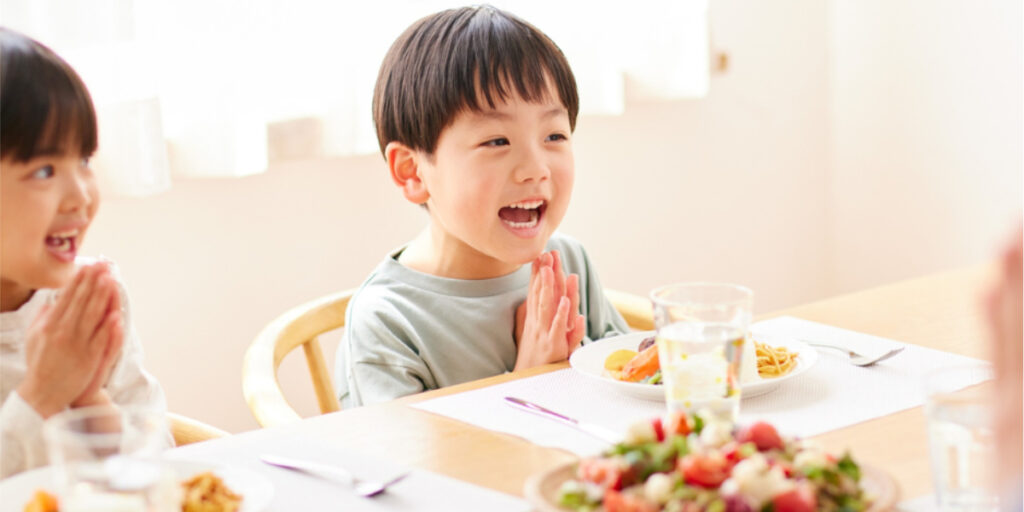 子どもたちが笑顔で「いただきます」をする食卓風景。健康料理家が提案する、食育とバランスの取れた家庭の食事シーン。